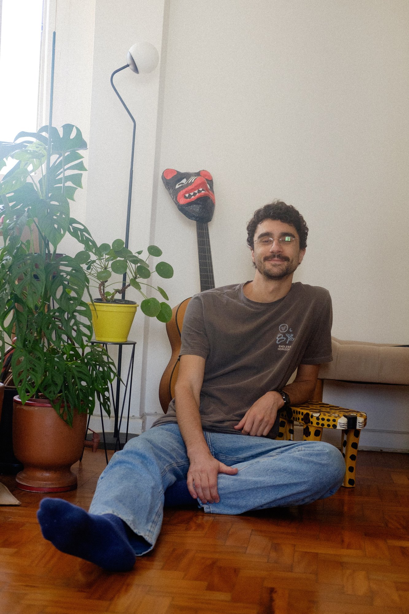 Artur sentado no chão da sala, de óculos, sorrindo, as pernas esticadas, diante das plantas e de um violão com uma máscara vermelha.