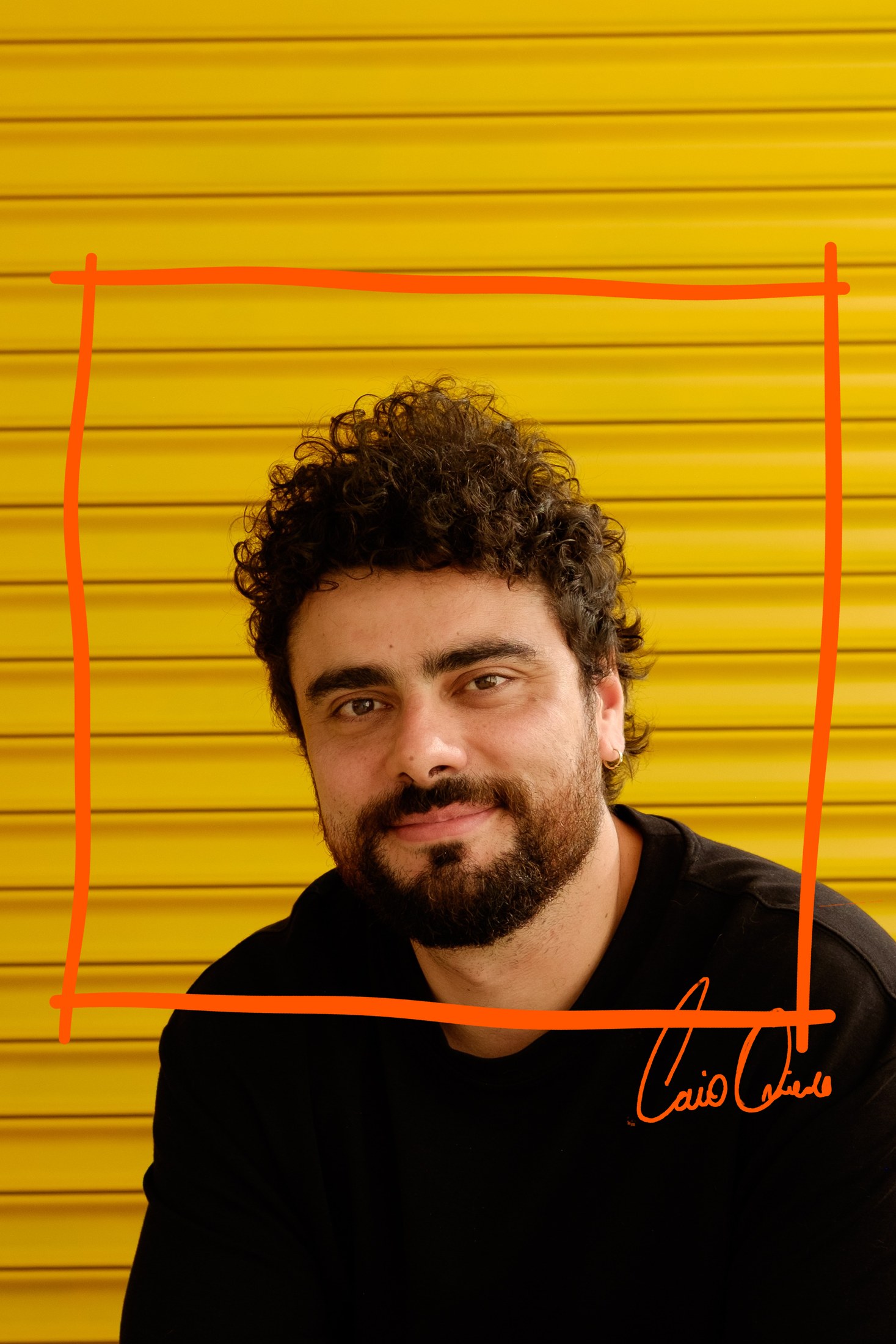 Caio, de cabelo cacheado e barba cheia, sorrindo de leve para a câmera diante de uma porta de enrolar amarela, de camiseta preta. Um quadrado laranja riscado à mão o envolve e a assinatura “Caio Oviedo” atravessa a quina de baixo, sobre a camiseta.