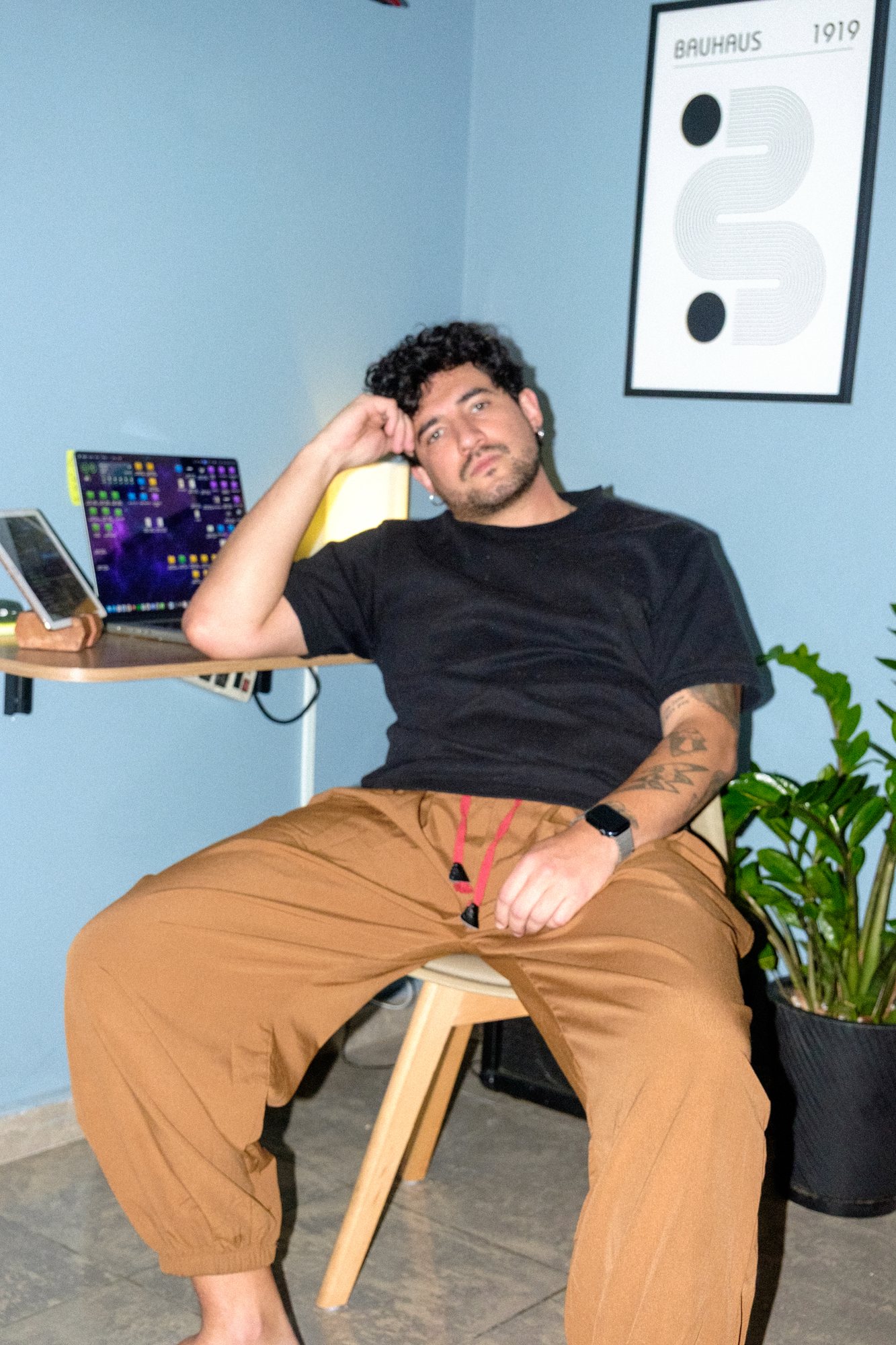 Wesley recostado numa cadeira de madeira, a mão na cabeça, diante da mesa com notebook e de um pôster da Bauhaus numa parede azul.
