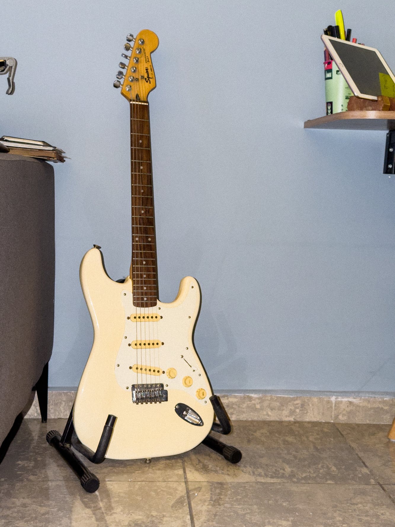Uma guitarra branca no suporte, encostada na parede azul, diante do sofá cinza; ao fundo, uma prateleira com um tablet.
