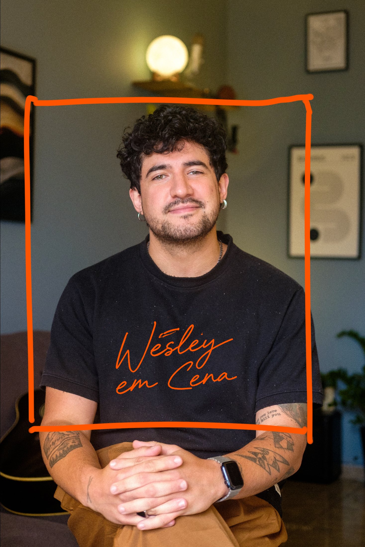 Wesley sentado diante de uma parede verde, de camiseta preta, barba e cabelo cacheado, as mãos entrelaçadas sobre o joelho. Um quadrado laranja riscado à mão o envolve e a assinatura “Wesley em Cena” cruza a camiseta.