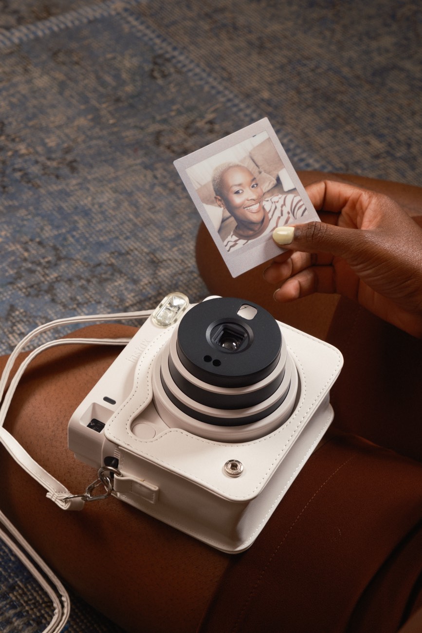 Mulher sorrindo com uma bolsa instax na mão, contra uma parede de madeira