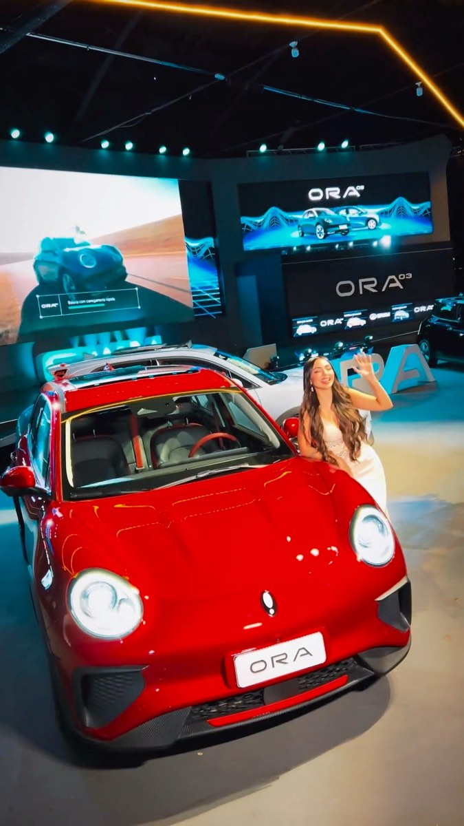 Carro vermelho no salão, com uma convidada ao lado e os telões atrás